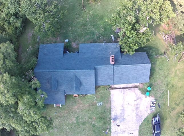 Aerial view of a dark-roofed house surrounded by trees, with a driveway and parked vehicle on the right.