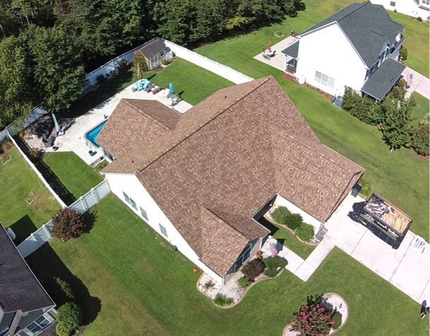 Aerial view of a suburban house with a brown roof, green lawn, driveway, and backyard patio.