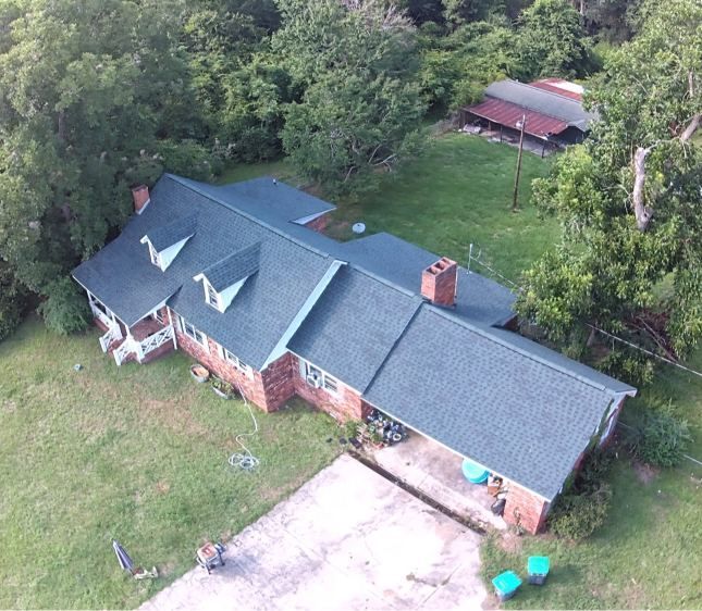 Aerial view of a red brick house with a gray roof, driveway, and wooded yard.