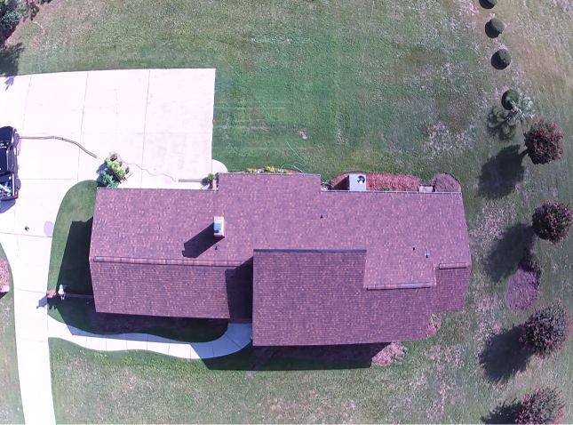 Aerial view of a house with a red roof, driveway, and grassy yard with scattered trees.
