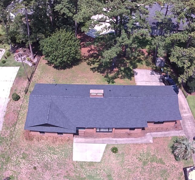 Aerial view of a single-story house with a dark roof, surrounded by a large grassy yard and trees.