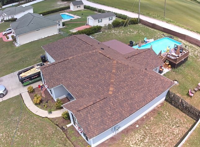 Aerial view of a house with a brown roof, driveway, yard, and backyard pool in a suburban neighborhood
