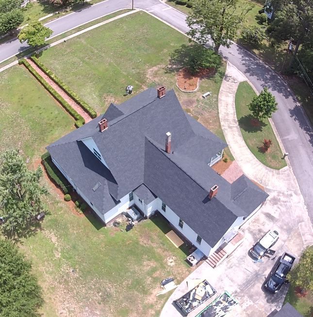 Aerial view of a gray-roofed house with driveway, lawn, and trees on a curved suburban street.