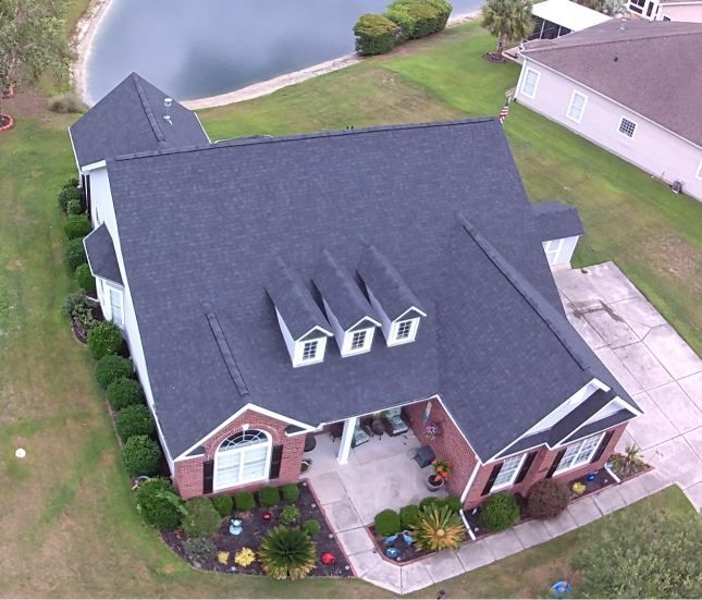 Aerial view of a brick house with a dark roof, front porch, and driveway in a suburban yard.