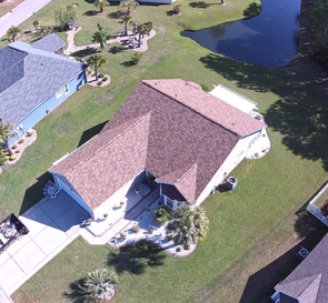 Aerial view of a suburban house with a brown roof, driveway, lawn, and a pond nearby.