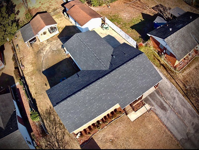 Aerial view of a house with a dark gray roof surrounded by brown yards and other houses.
