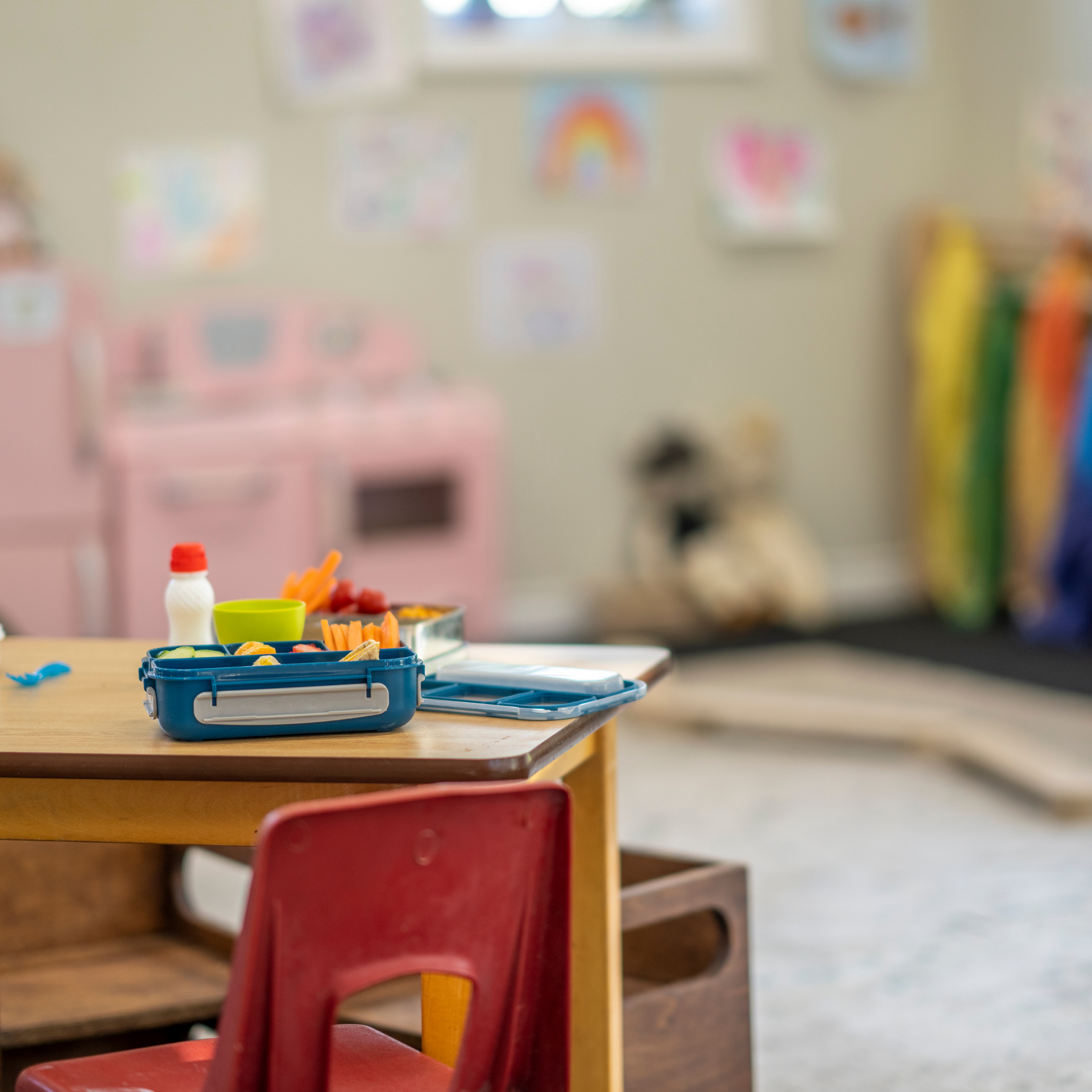 Lunchbox on a table in a child's playroom; red chair in the foreground, toy kitchen and artwork in the background.