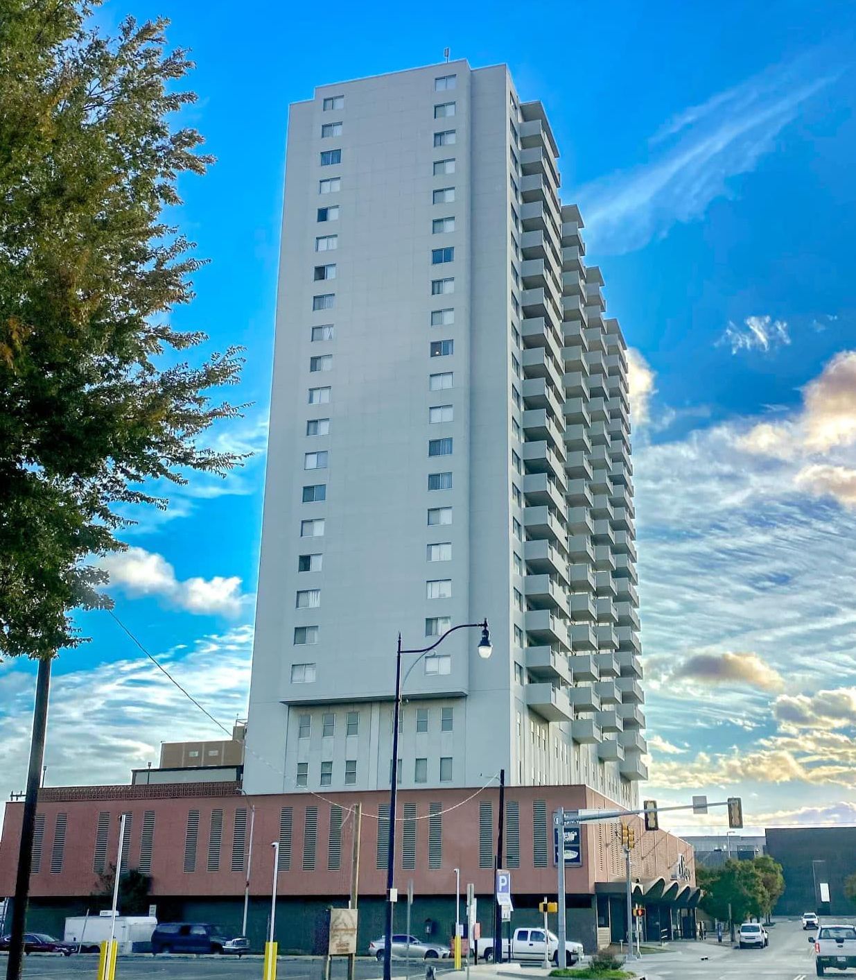 Tall light gray high-rise apartment building with balconies and a lower pink brick section, set against a blue sky.