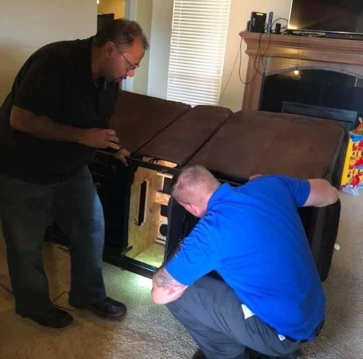 Two men assembling a brown couch in a living room. One man stands, the other crouches, both focused on the furniture.