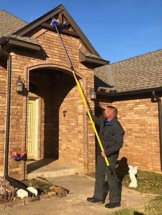 Man using a yellow extended pole with a blue brush to clean a house’s exterior under a clear sky.