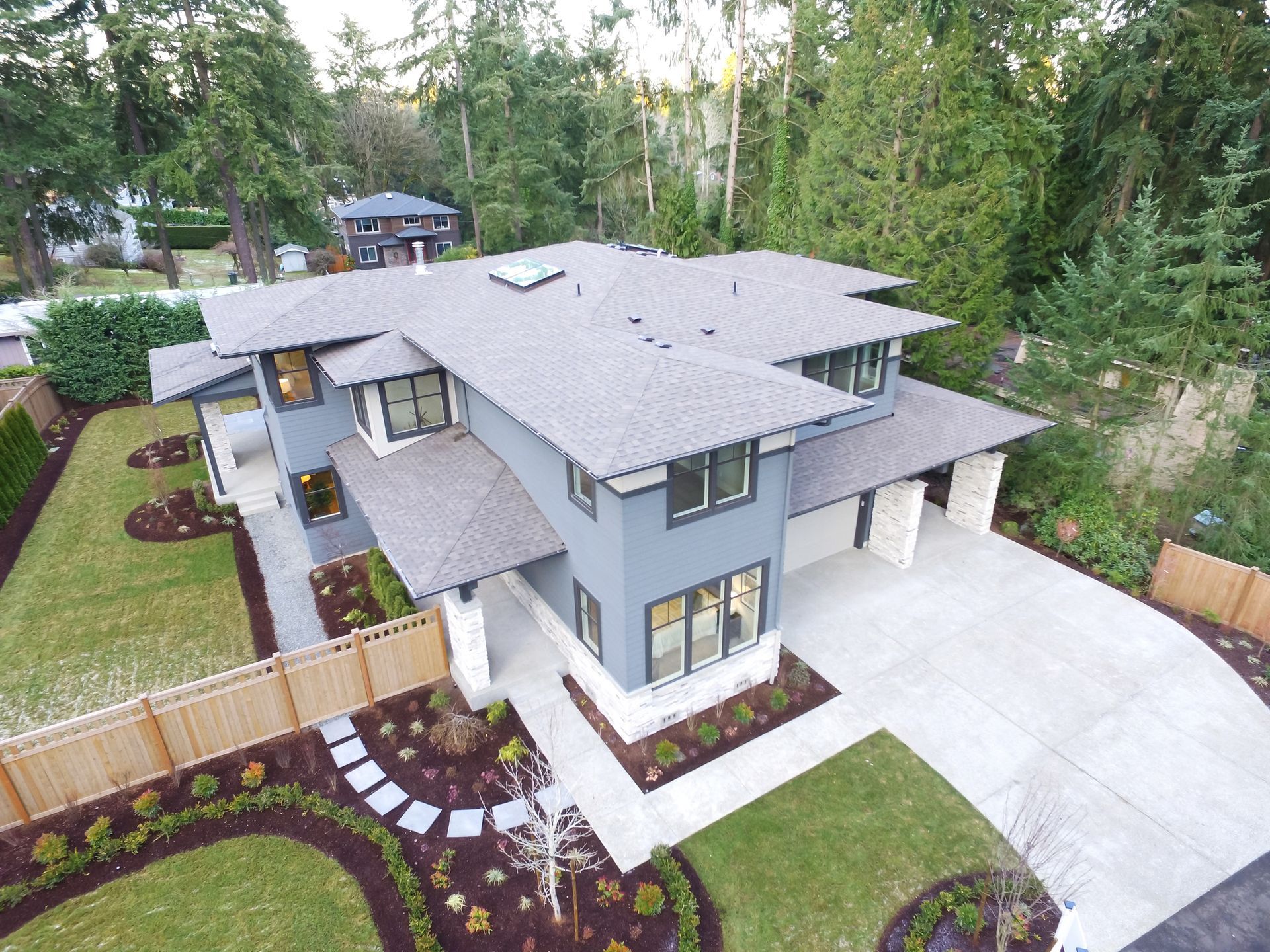 Modern two-story gray house with a concrete driveway, landscaped yard, and surrounding trees.