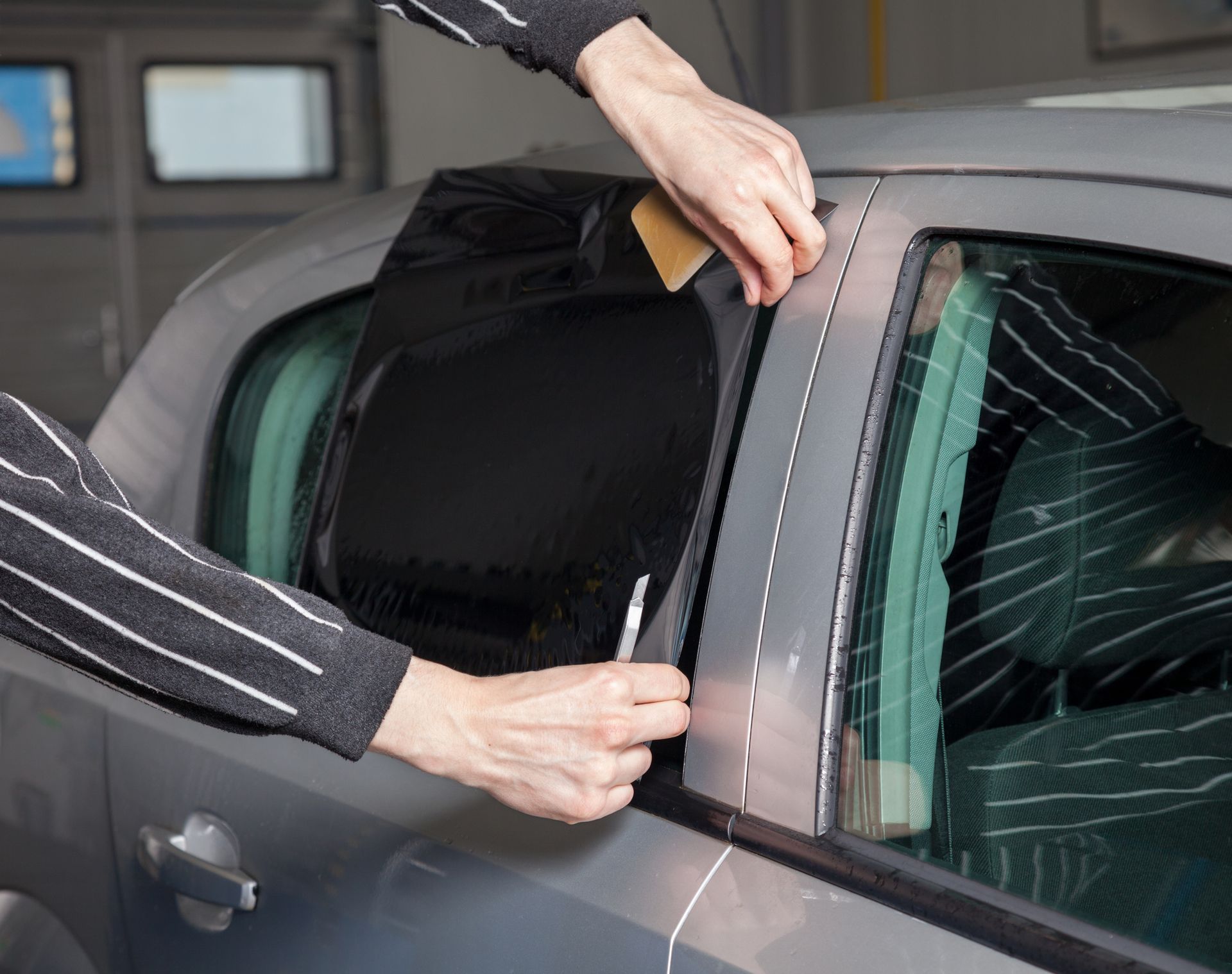 A person applying window tint to a gray car window with tools inside a garage.