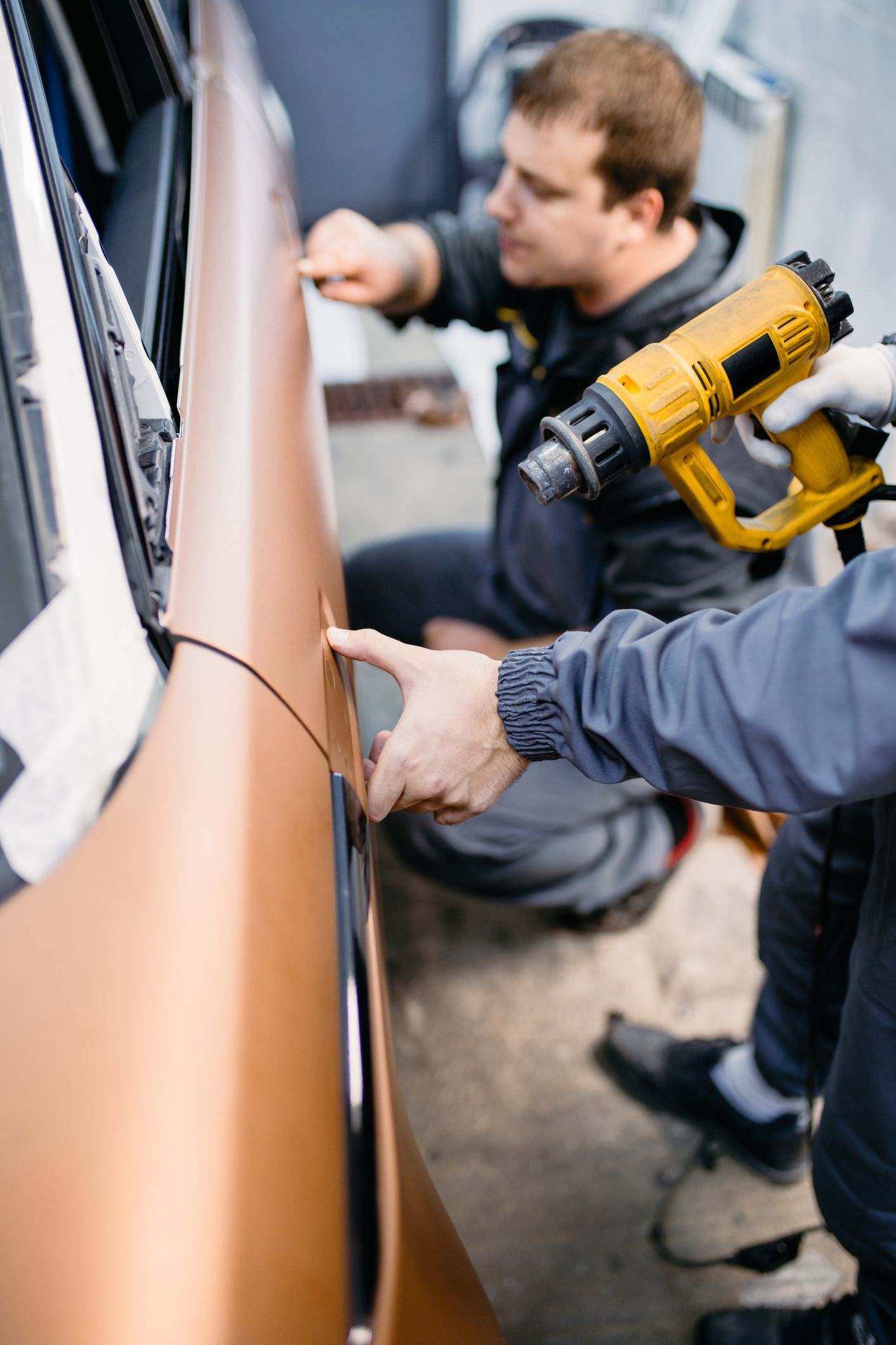 Two people applying orange vinyl wrap to a car door, one using a heat gun.