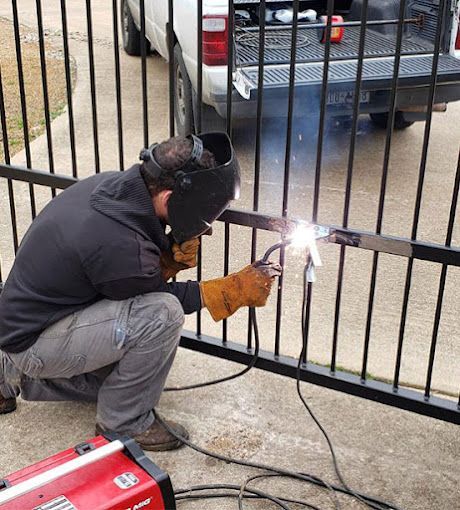 Worker welding a black metal gate outdoors, wearing a helmet and gloves, with sparks flying.