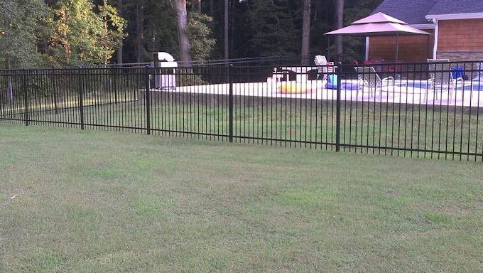 Black metal fence around a grassy yard with a pool, umbrellas, and a small pavilion in the background