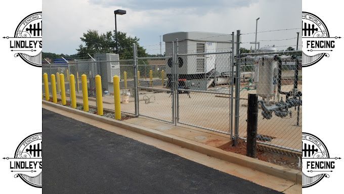 Industrial fenced utility area with chain-link gate, yellow bollards, and gray equipment on a paved lot