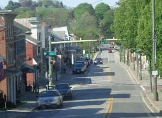 Main street with parked cars, storefronts, trees, and a few moving vehicles on a sunny day Main street with parked cars, storefronts, trees, and a few moving vehicles on a sunny day