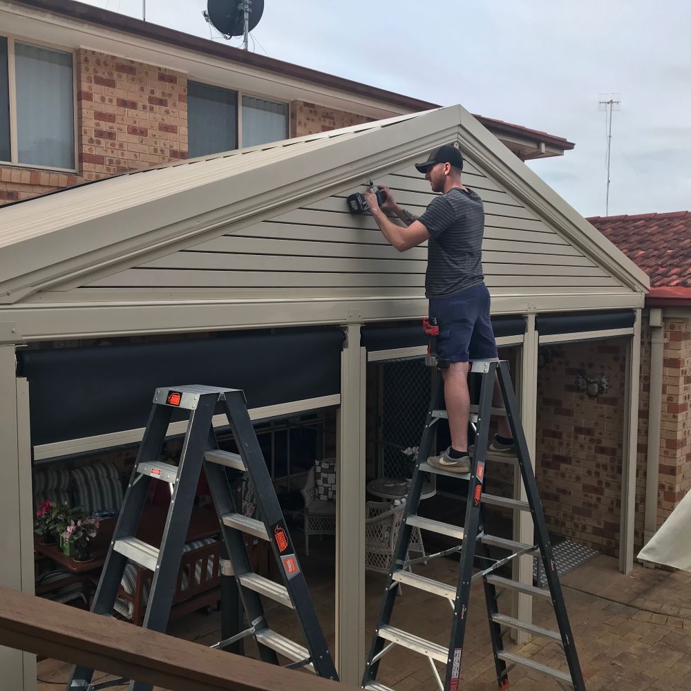 A Man is Standing on a Ladder Working on a Building — Outdoor Leisure Living In Tuggerah, NSW