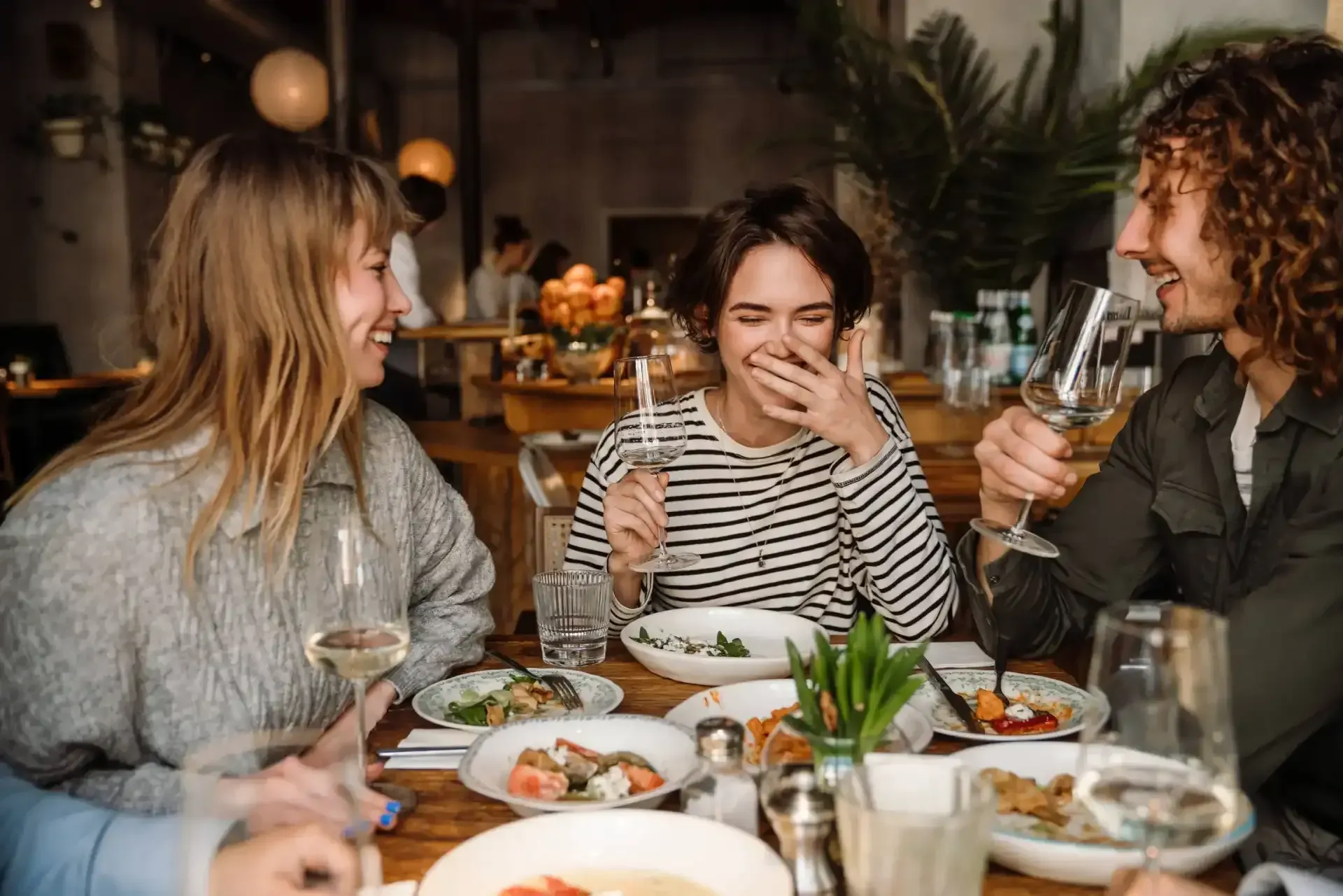 Three people laughing and drinking wine at a restaurant table with food, plants, and natural light.