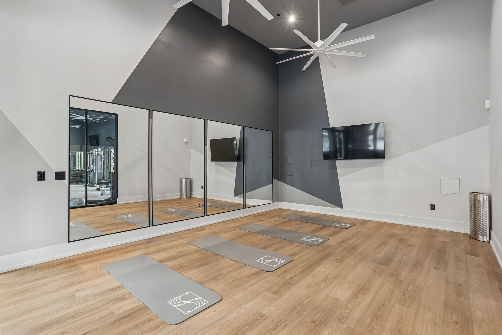 A modern yoga or fitness studio with wooden floors, grey yoga mats, a mirrored wall, and geometric grey wall accents.