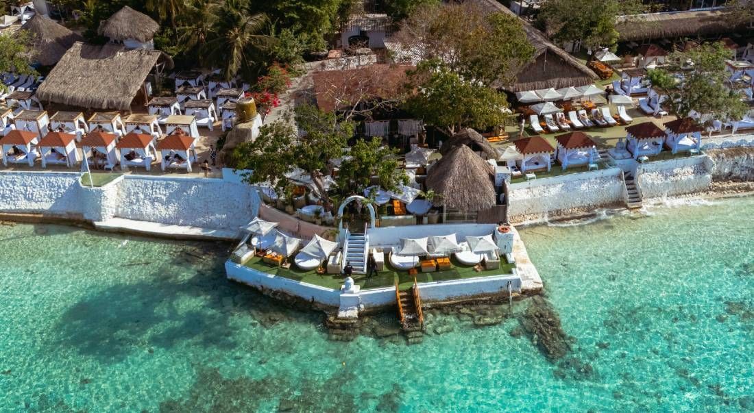 Vista aérea de un resort frente a la playa con cabañas blancas, agua turquesa y techos de paja.  | Mevak Agency