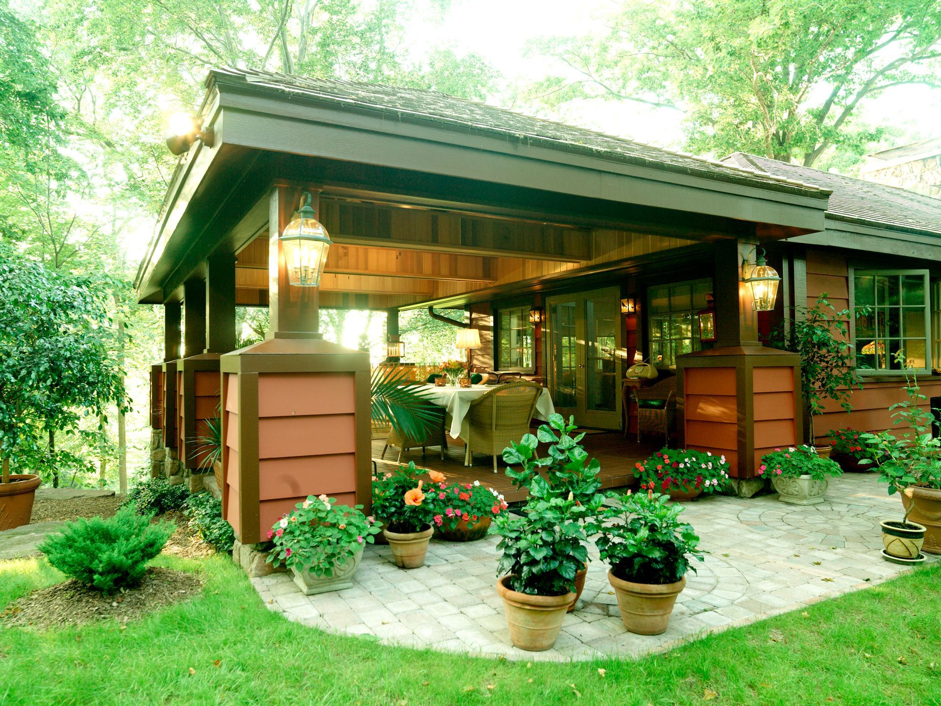 Covered patio with potted plants on a brick patio and a lawn, next to a home.