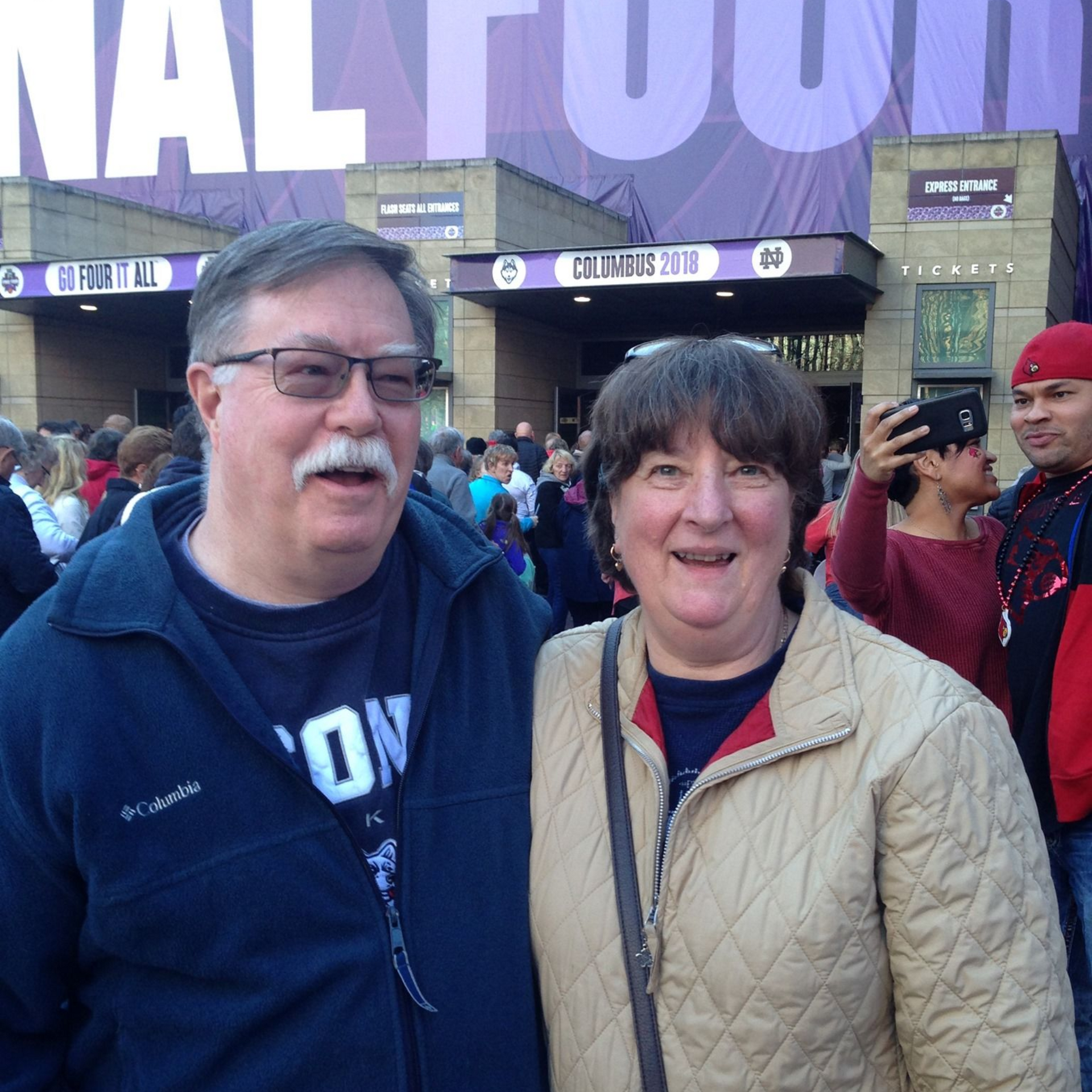 A smiling man and woman pose for a photo in front of a stadium entrance marked