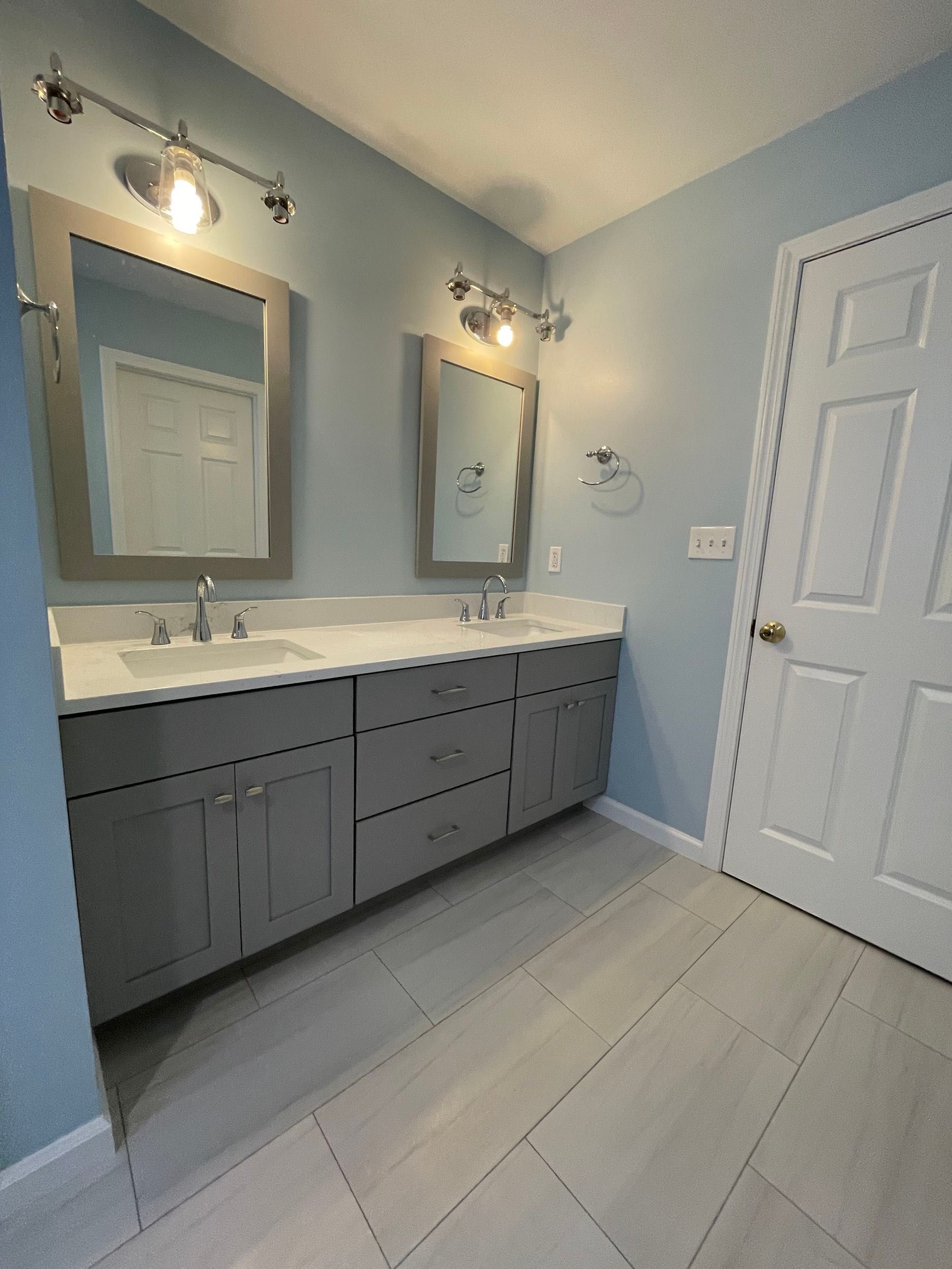 Bathroom with gray double vanity, light blue walls, two mirrors, and a white door.