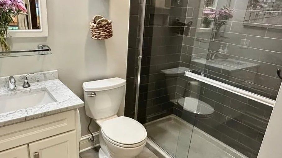 Bathroom with toilet, sink, and glass-enclosed shower. Gray and white color scheme, basket on wall.