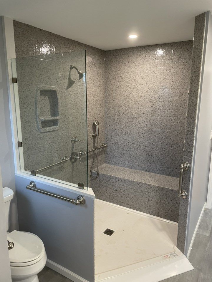 Accessible shower with grab bars, bench, and glass door. Gray mosaic tile, white floor, and chrome fixtures.