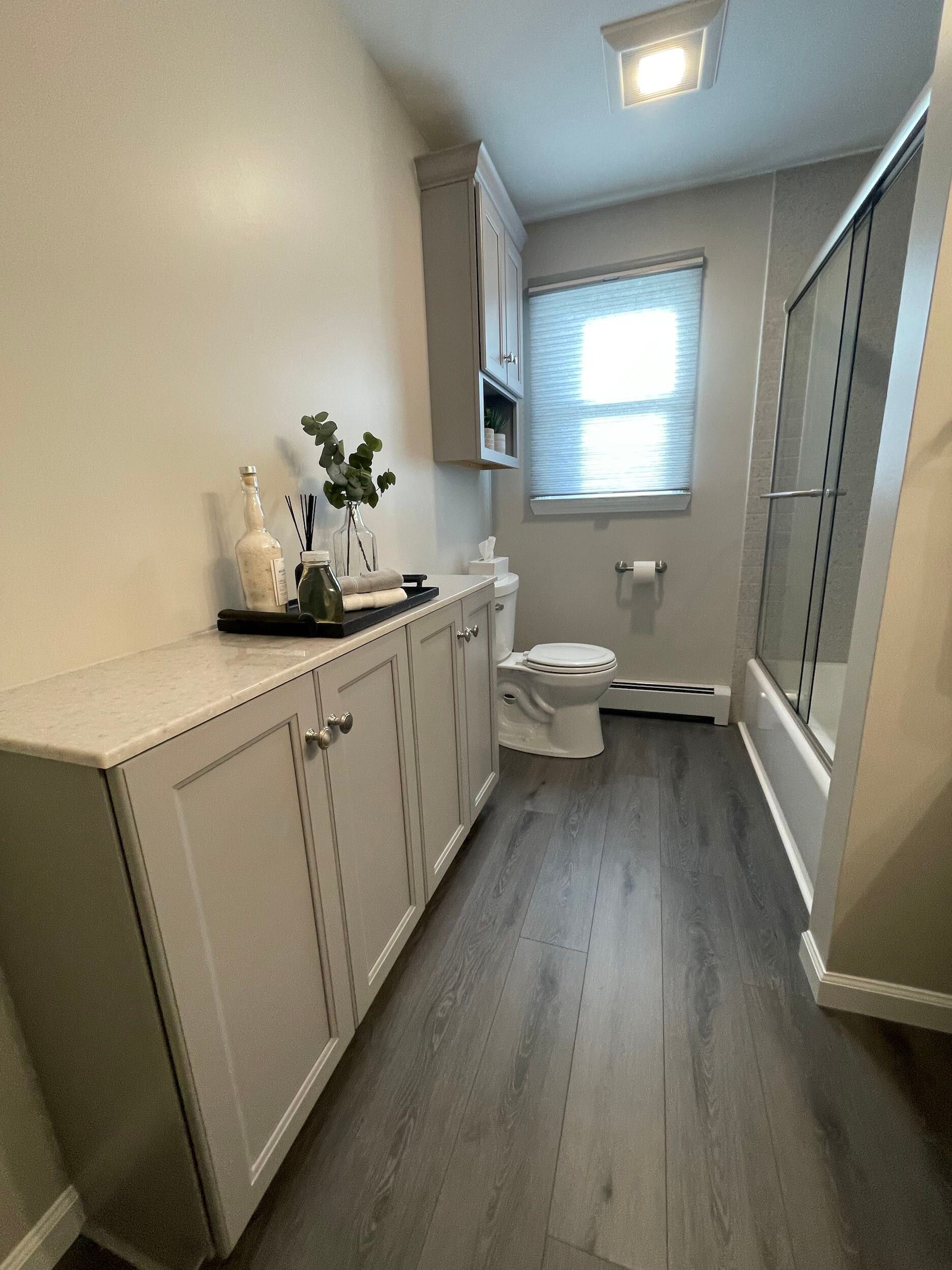 Bathroom with gray cabinets, gray flooring, toilet, shower, and window.