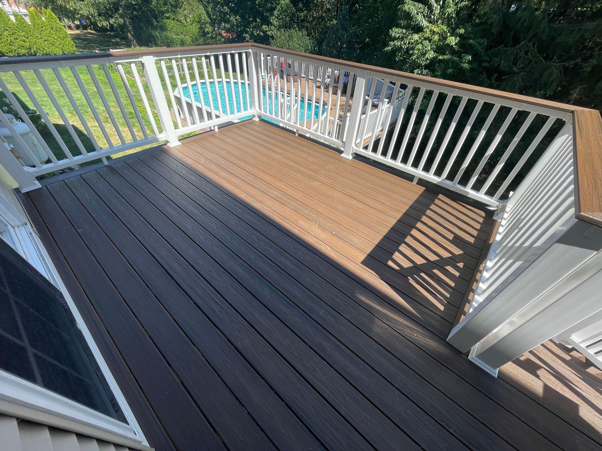 Wooden deck with white railing, overlooking a pool. Sunlight casts shadows on the deck.