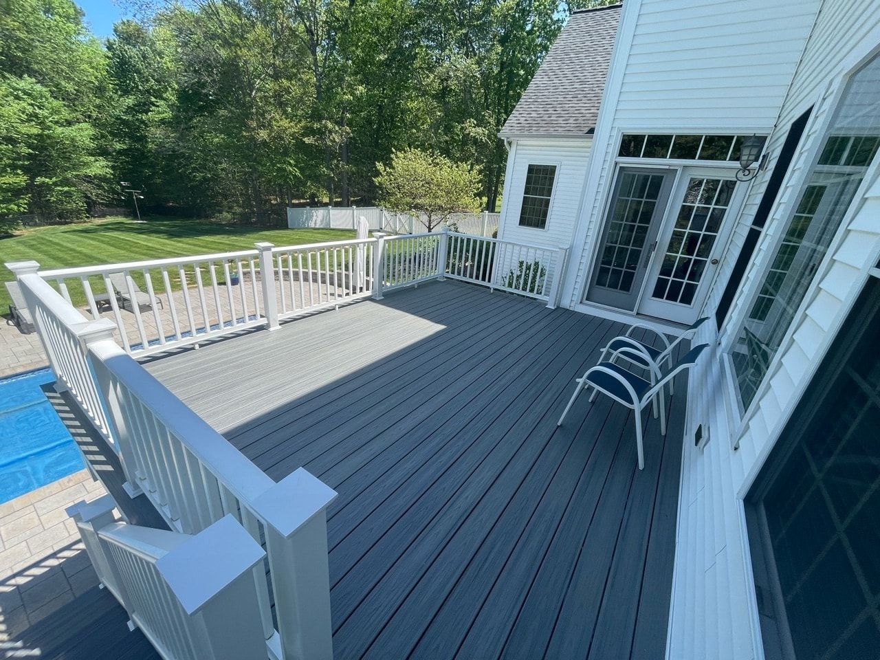 Gray deck with white railing next to a pool and white house with glass doors. Sunny day.