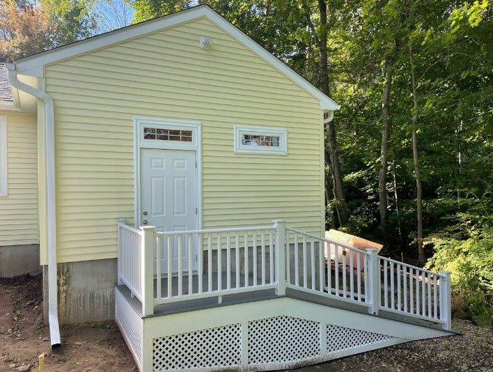 Yellow building with white door, windows, and ramp with railing. Overlooking trees.
