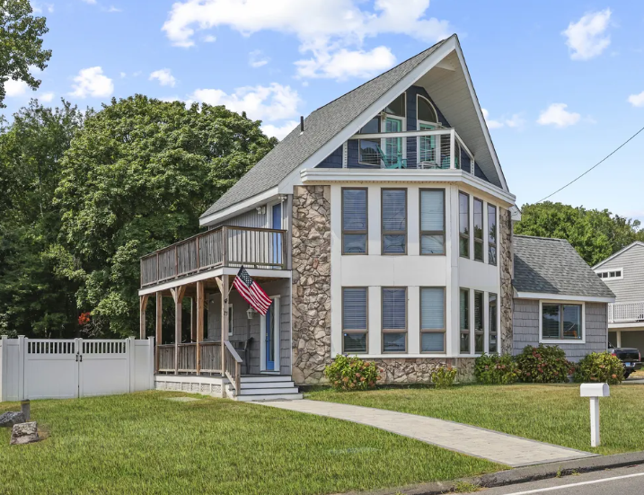 Two-story house with stone facade, wrap-around porch, and American flag; green lawn, sunny day.
