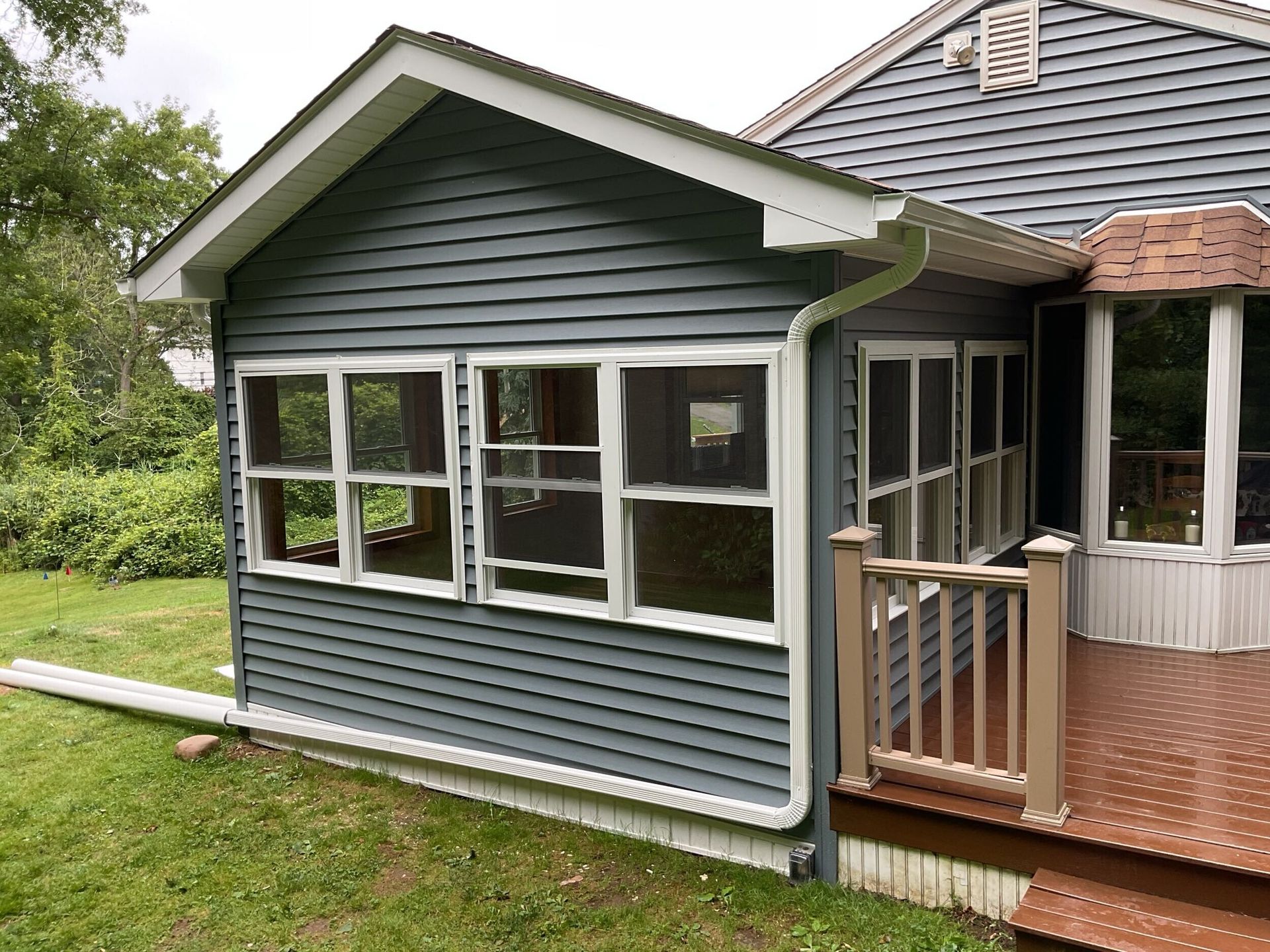 Sunroom addition with gray siding and white-framed windows, attached to a house with a wooden deck.