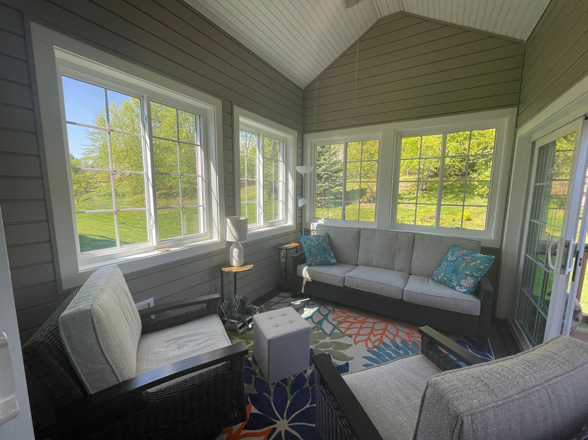 Sunroom with gray walls, white-framed windows, wicker furniture, and a colorful rug, overlooking greenery.