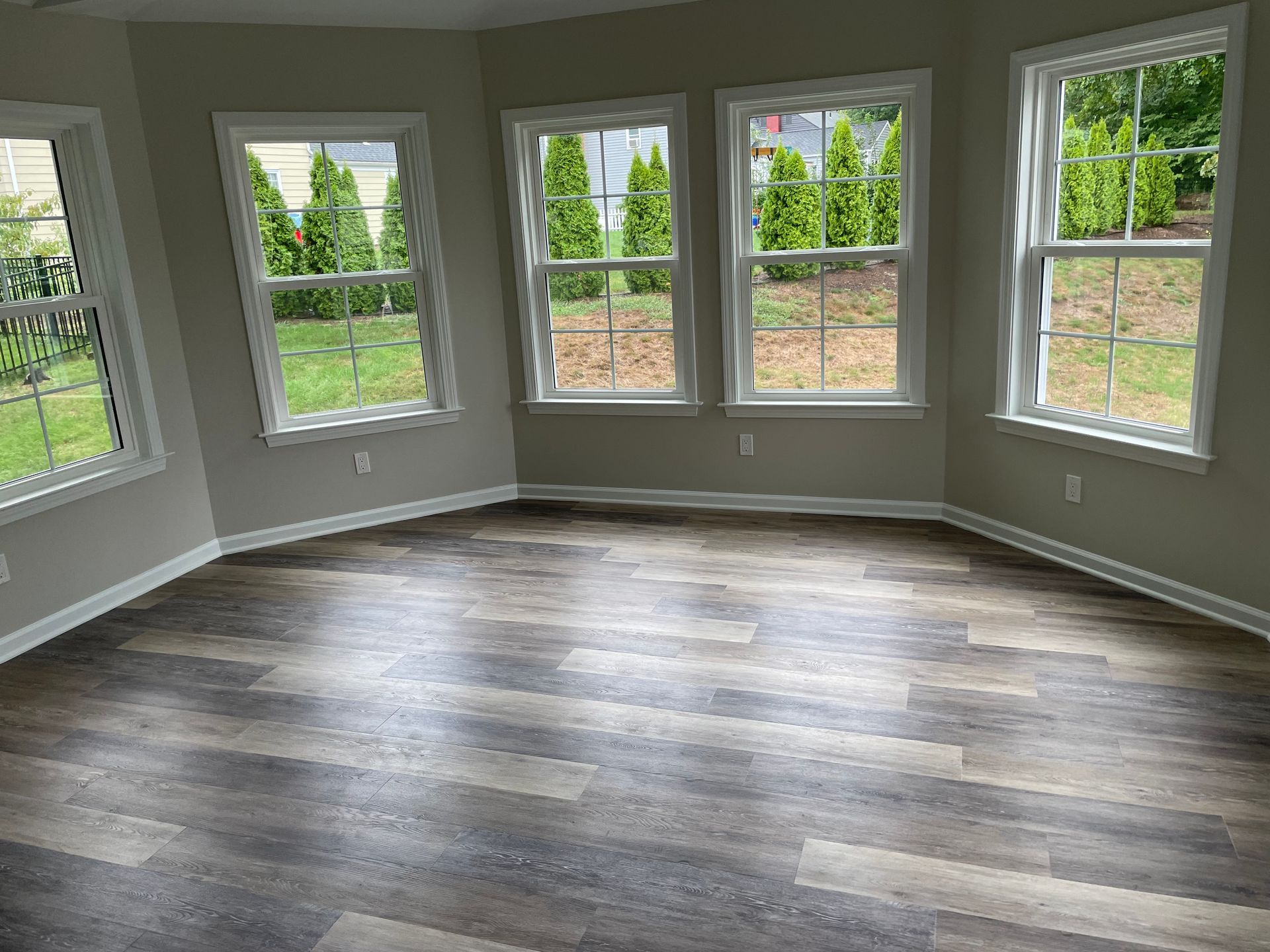 Empty room with gray-brown wood-look flooring, multiple windows with outside greenery, and light gray walls.