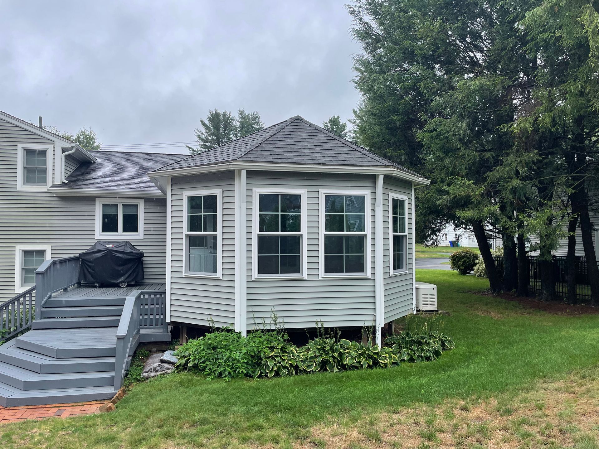 Backyard with a gray, octagonal sunroom and a deck with stairs, under a cloudy sky.