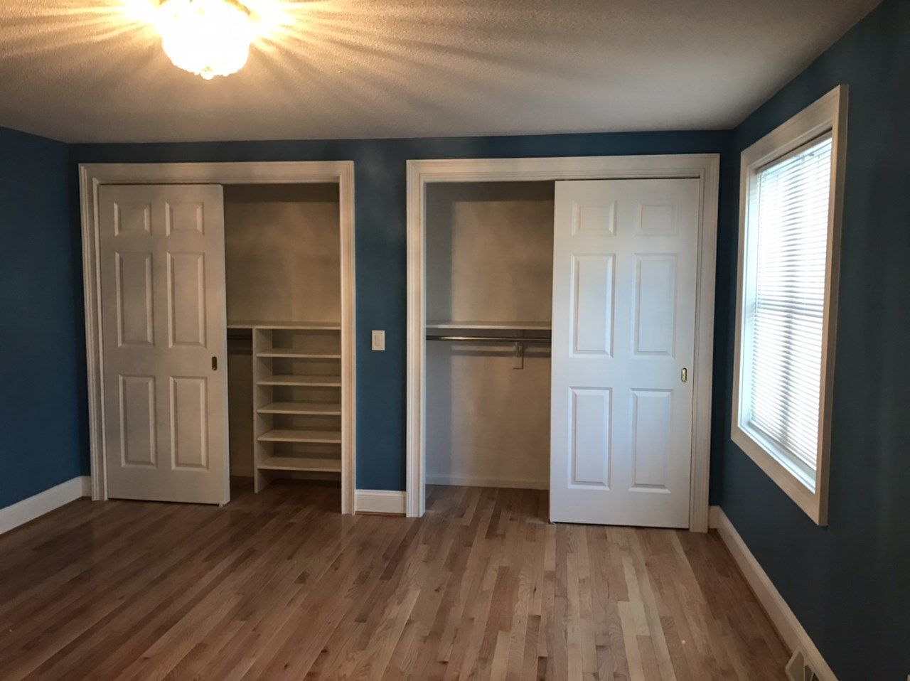 Bedroom with blue walls, hardwood floors, two closets with white doors, and a window with blinds.