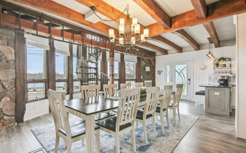Dining room with long table, chandelier, stone wall, and windows overlooking water.