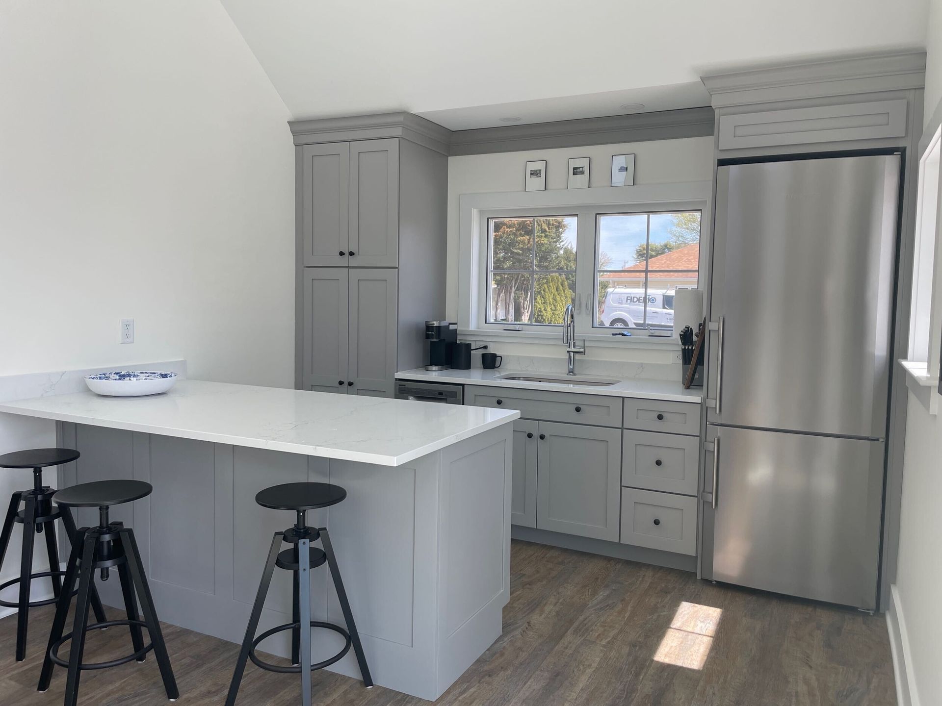 Gray kitchen with island, cabinets, and stainless steel fridge. Bar stools at the island. Light wood floors.