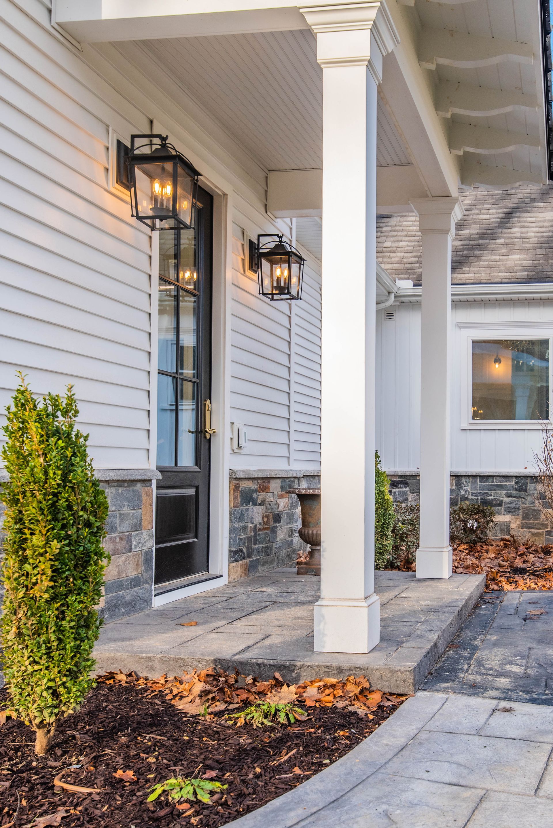 White home with a covered porch, black door, and lantern lights. Stone and concrete pathway, and small bush.