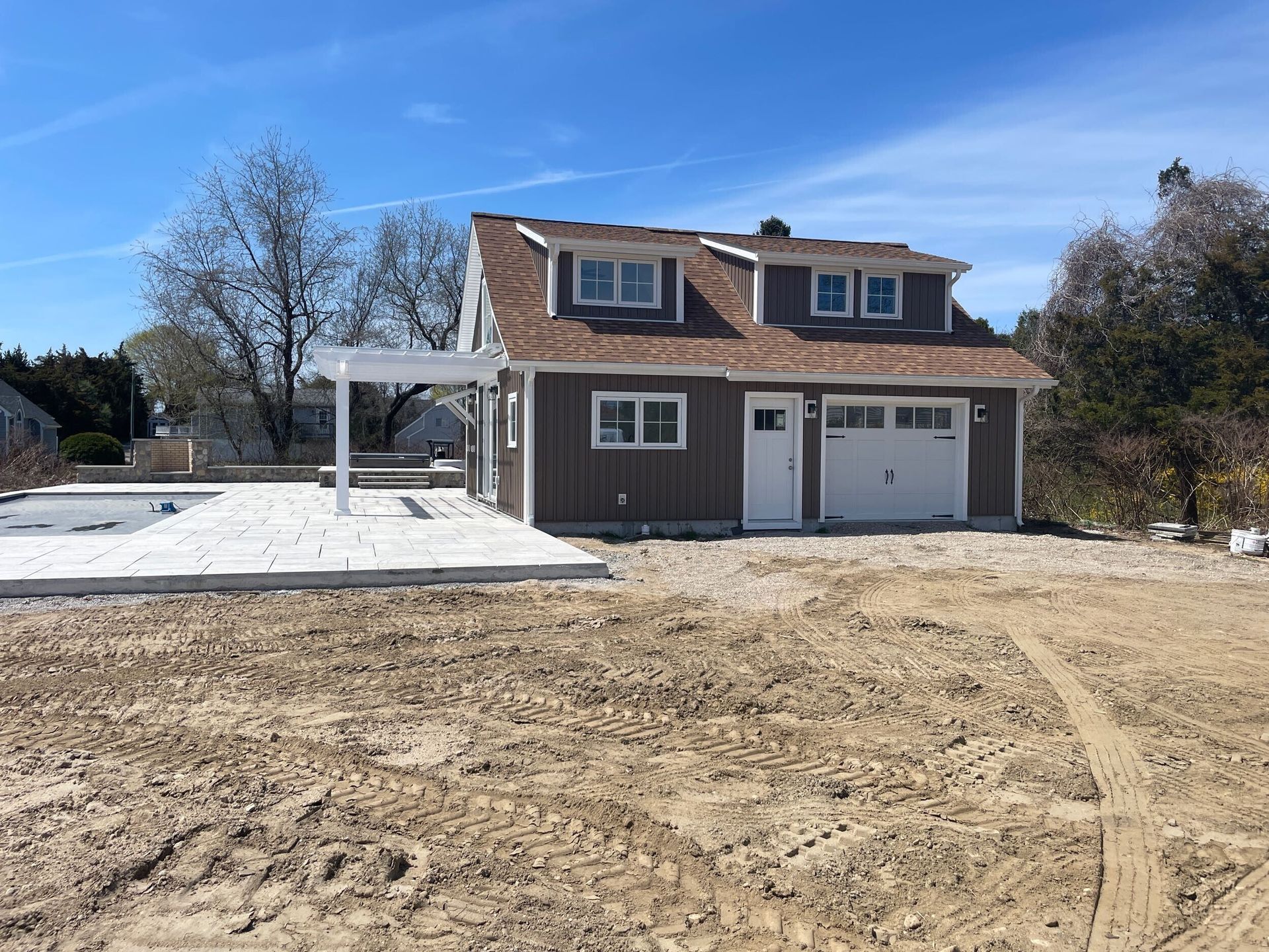 Brown house with white garage door and pergola, on a gravel lot under a blue sky.