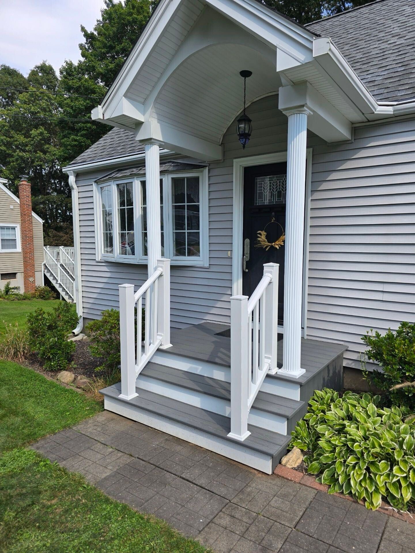 Gray house with white porch steps, railings, and columns; a black door with a gold decoration; and a green lawn.