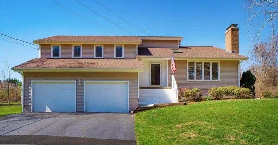 Two-story tan house with a two-car garage, a green lawn, and a blue sky.
