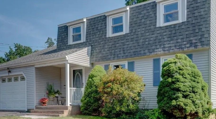 Two-story house with gray roof, white siding, blue shutters, and green bushes in front.