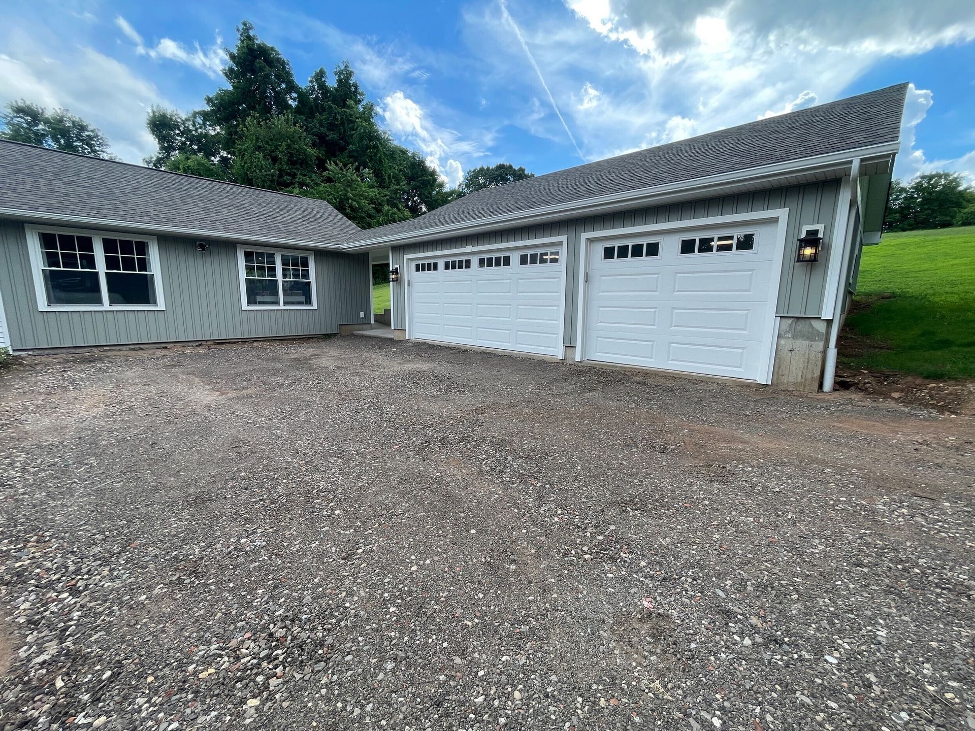A light gray house with two white garage doors and gravel driveway on a sunny day.