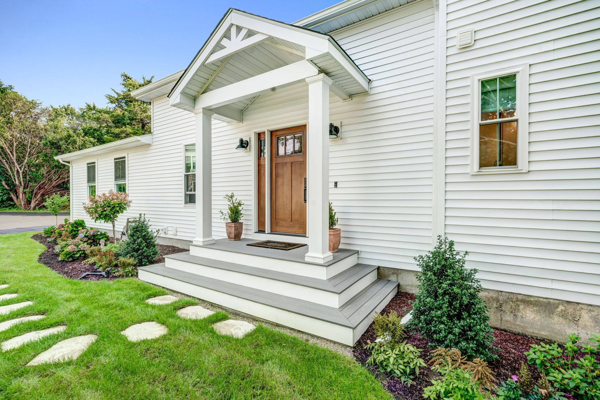 White house with wooden door and porch steps; green lawn with stepping stones.