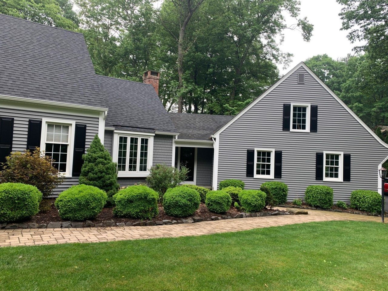 Gray house with dark shutters, trimmed bushes, brick pathway, and green lawn.